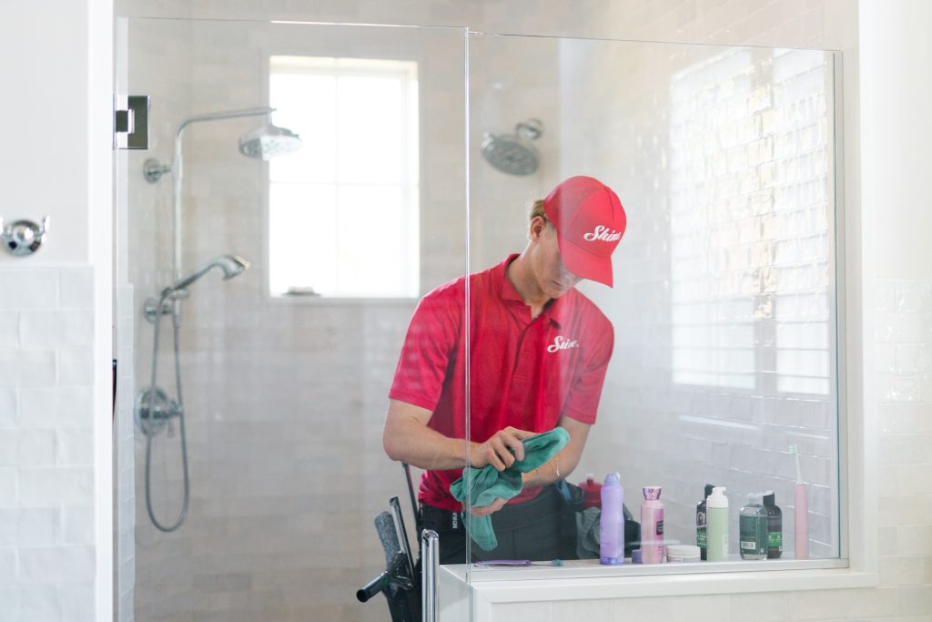 A cleaner wearing a red uniform meticulously cleans a glass shower enclosure.