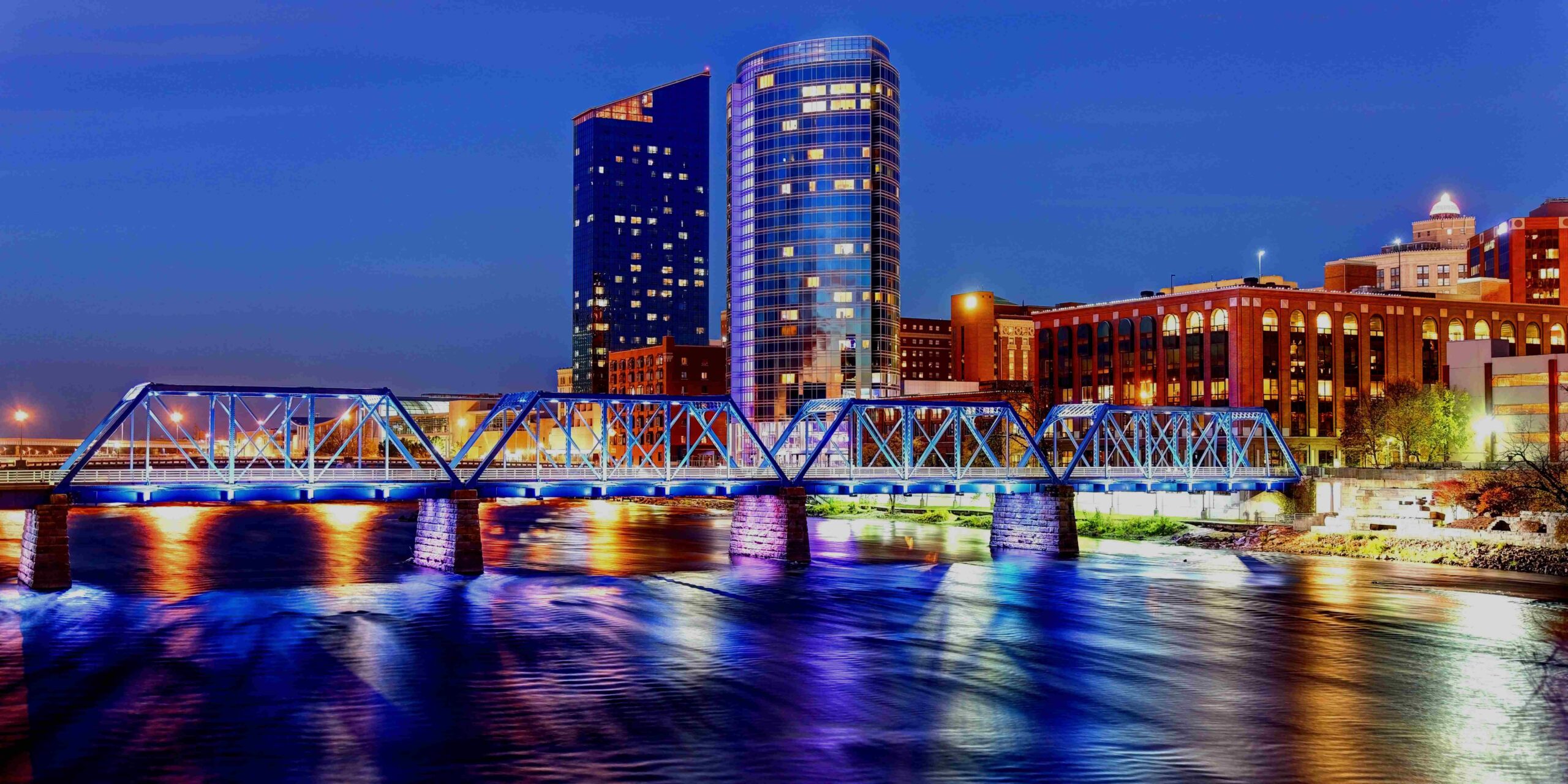A blue illuminated truss bridge spans a river reflecting city lights and a modern skyline at
