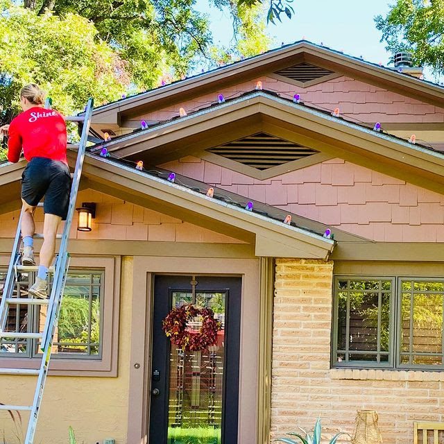 A person on a ladder installs colorful decorative lights along a house's roofline