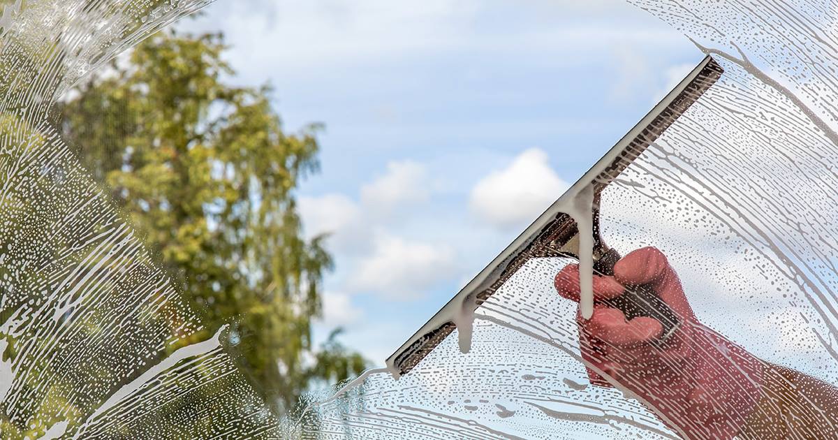 A hand uses a squeegee to clean a soapy window, revealing a clear view