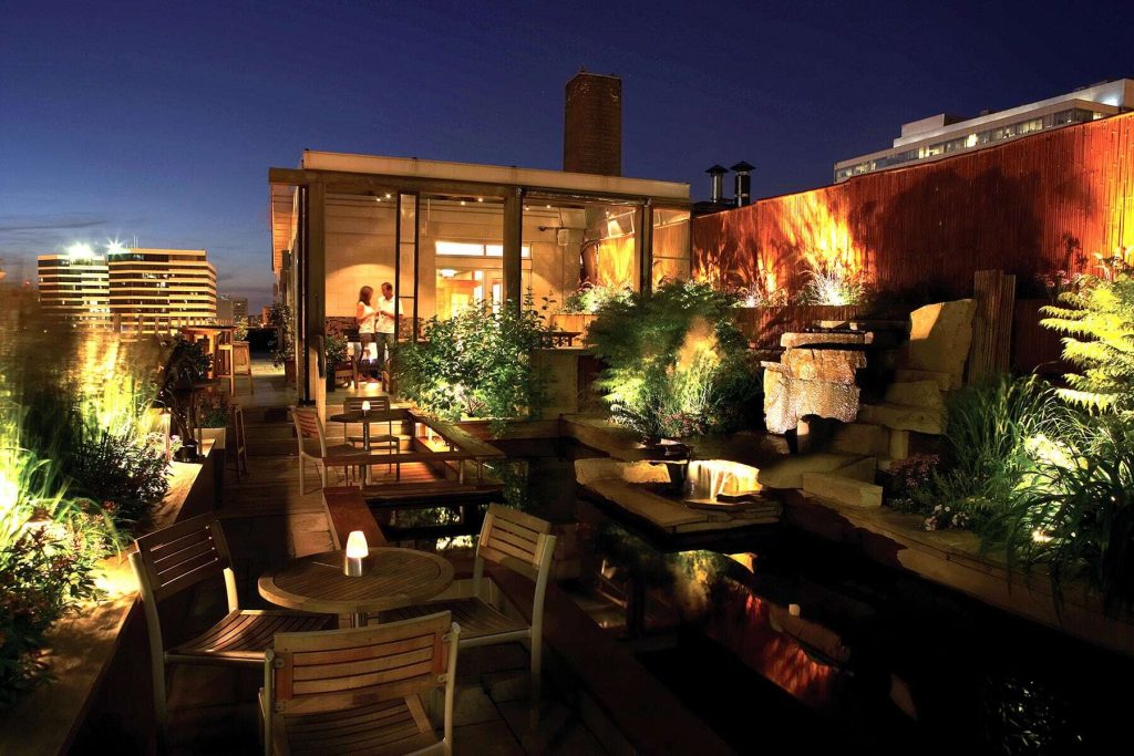 A beautifully lit rooftop patio at night features a pond, lush plants, and dining tables.
