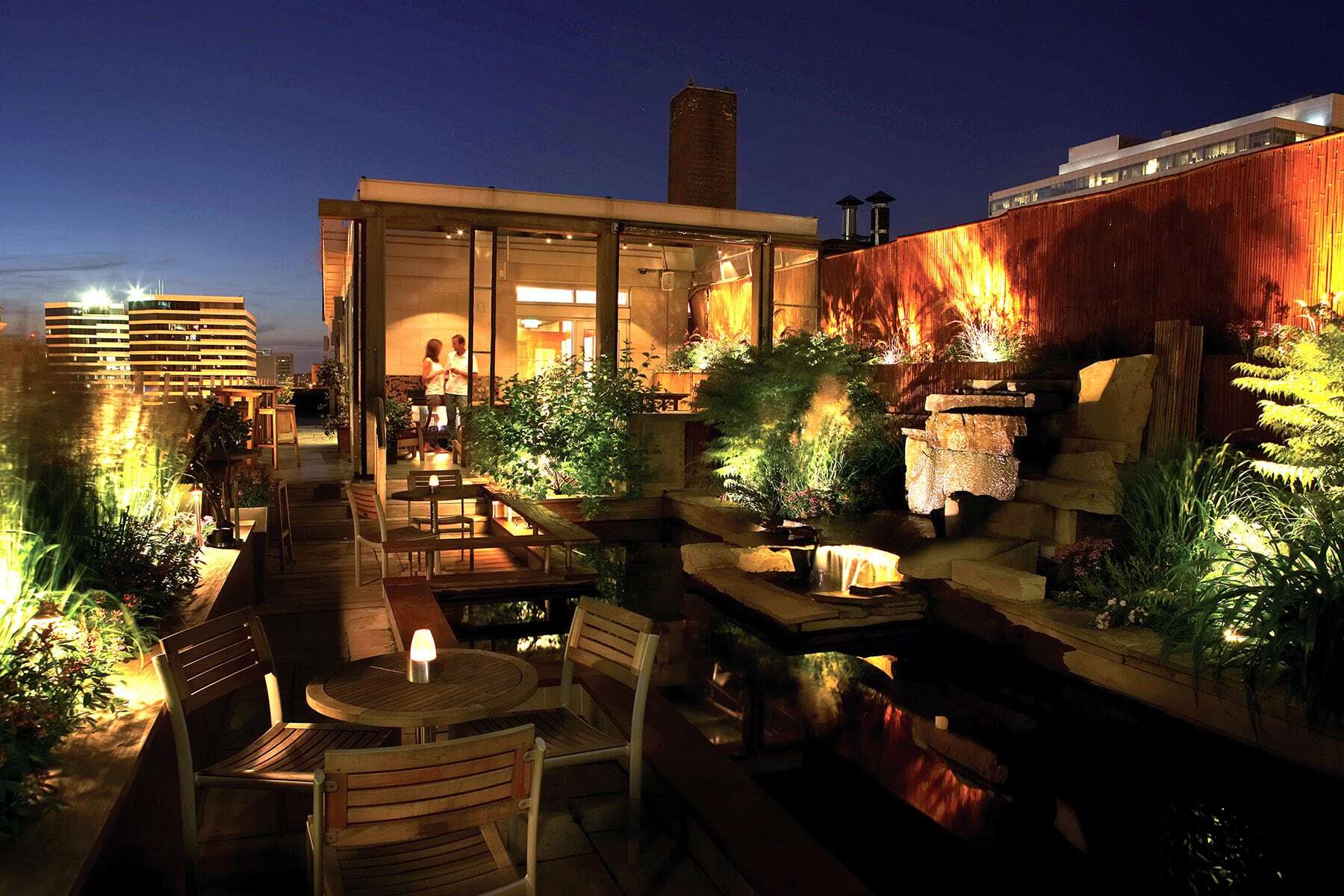 A beautifully lit rooftop patio at night features a pond, lush plants, and dining tables.