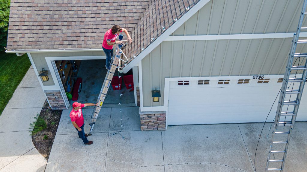 Two workers install string lights on a house roof from a ladder, with another worker spotting below.
