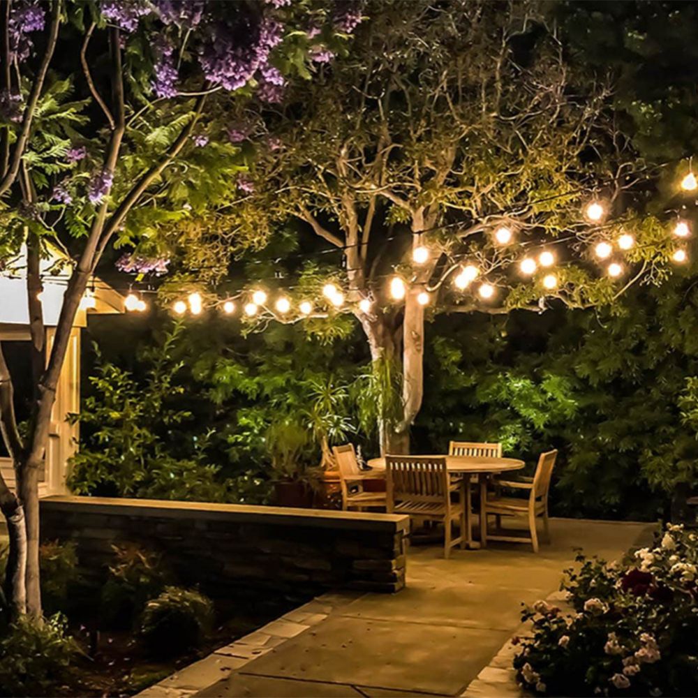 A cozy outdoor patio at night with string lights illuminating a wooden table and chairs surrounded by lush trees