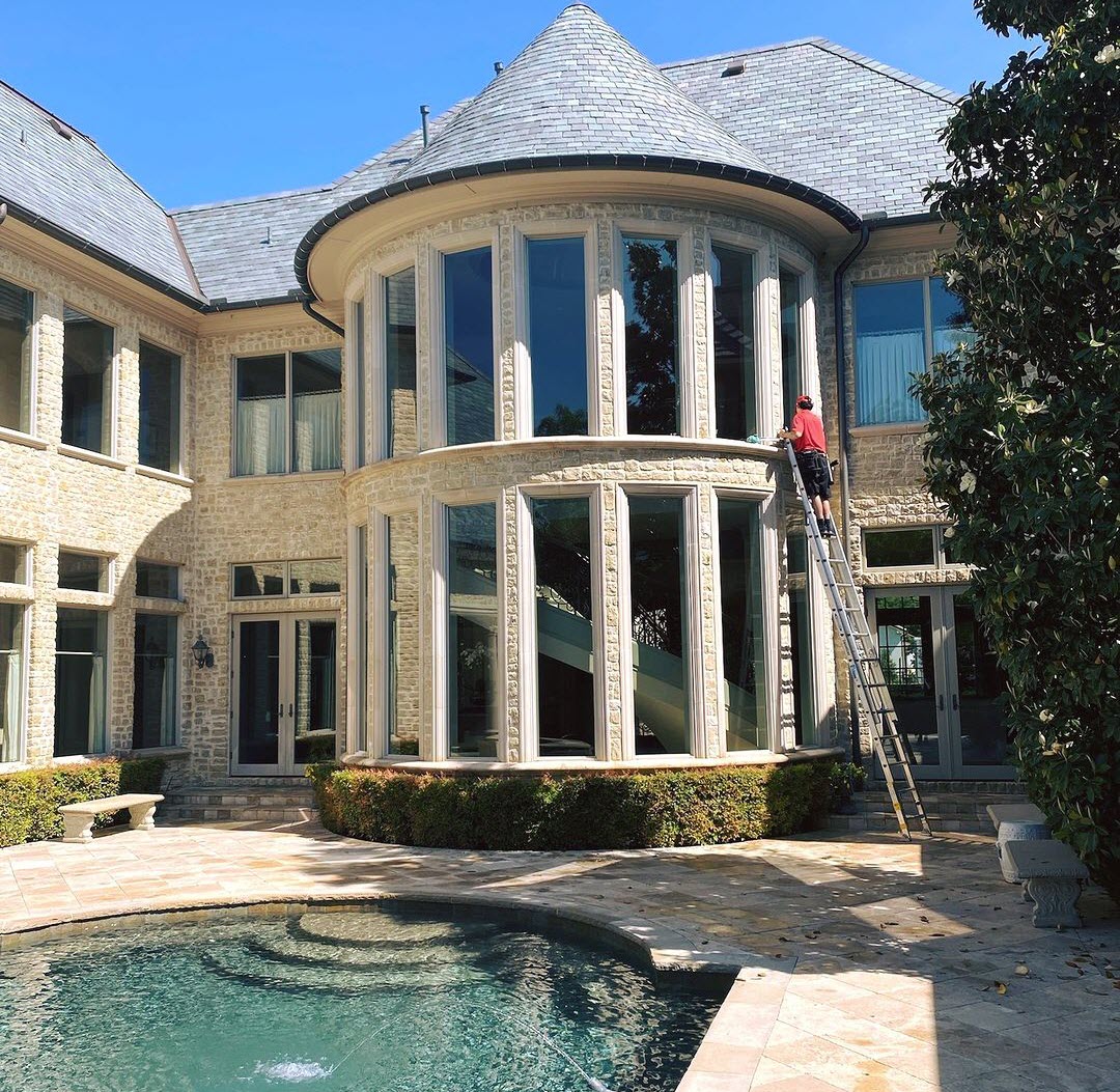 A worker on a ladder cleans the many windows of a grand stone mansion with a swimming
