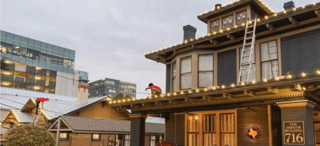 Workers install warm string lights on a historic building and adjacent structures in an urban setting.