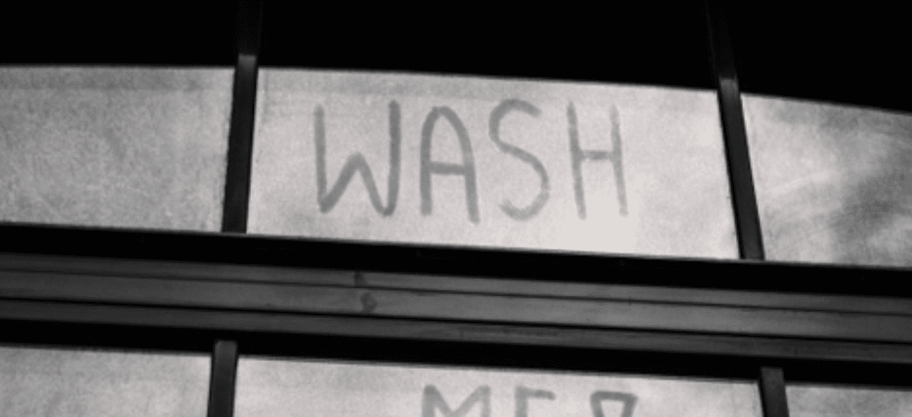 The words "WASH ME" are scrawled on a dirty, grimy window in