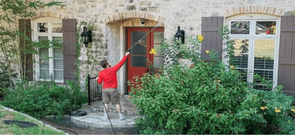 A man cleans the arched red doorway of a stone house with a long-reach pole.