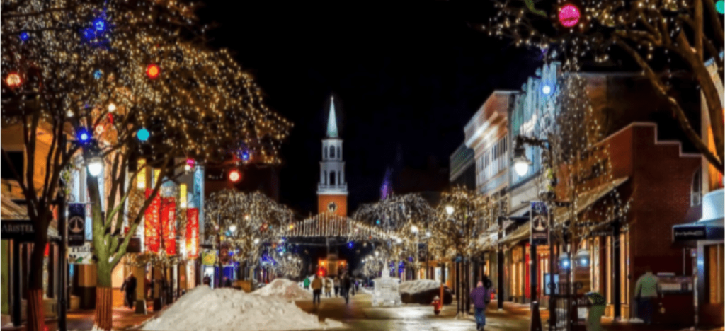 A vibrant downtown street at night, decorated with twinkling holiday lights, snow, and a