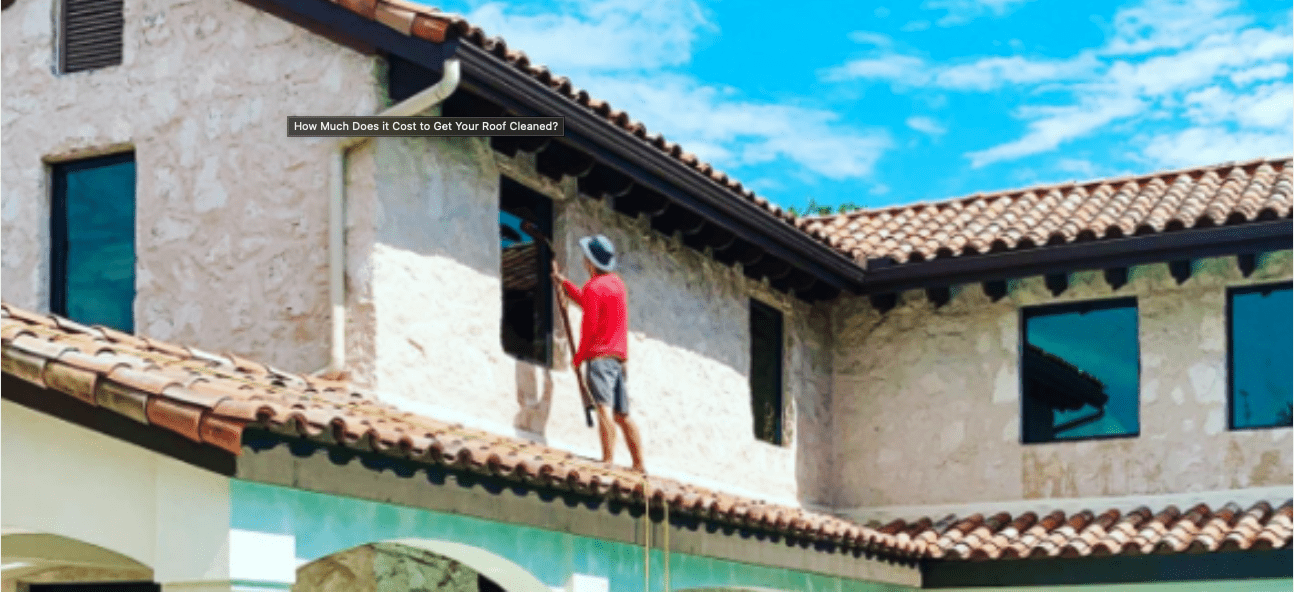 A worker stands on a tiled roof, cleaning a window of a stucco building under