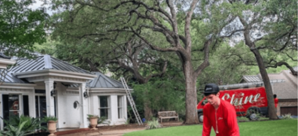 A worker in a red uniform stands on a lawn outside a white house with a red 'Sh