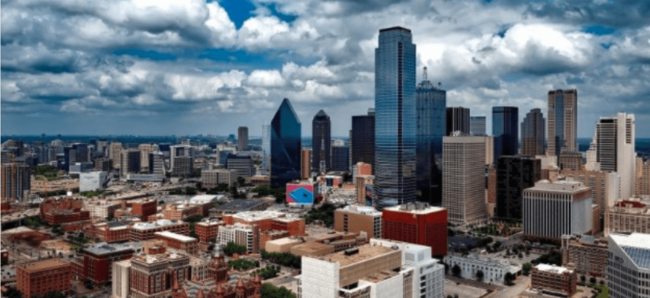 The Dallas city skyline features modern skyscrapers and lower buildings under a dramatic cloudy sky.