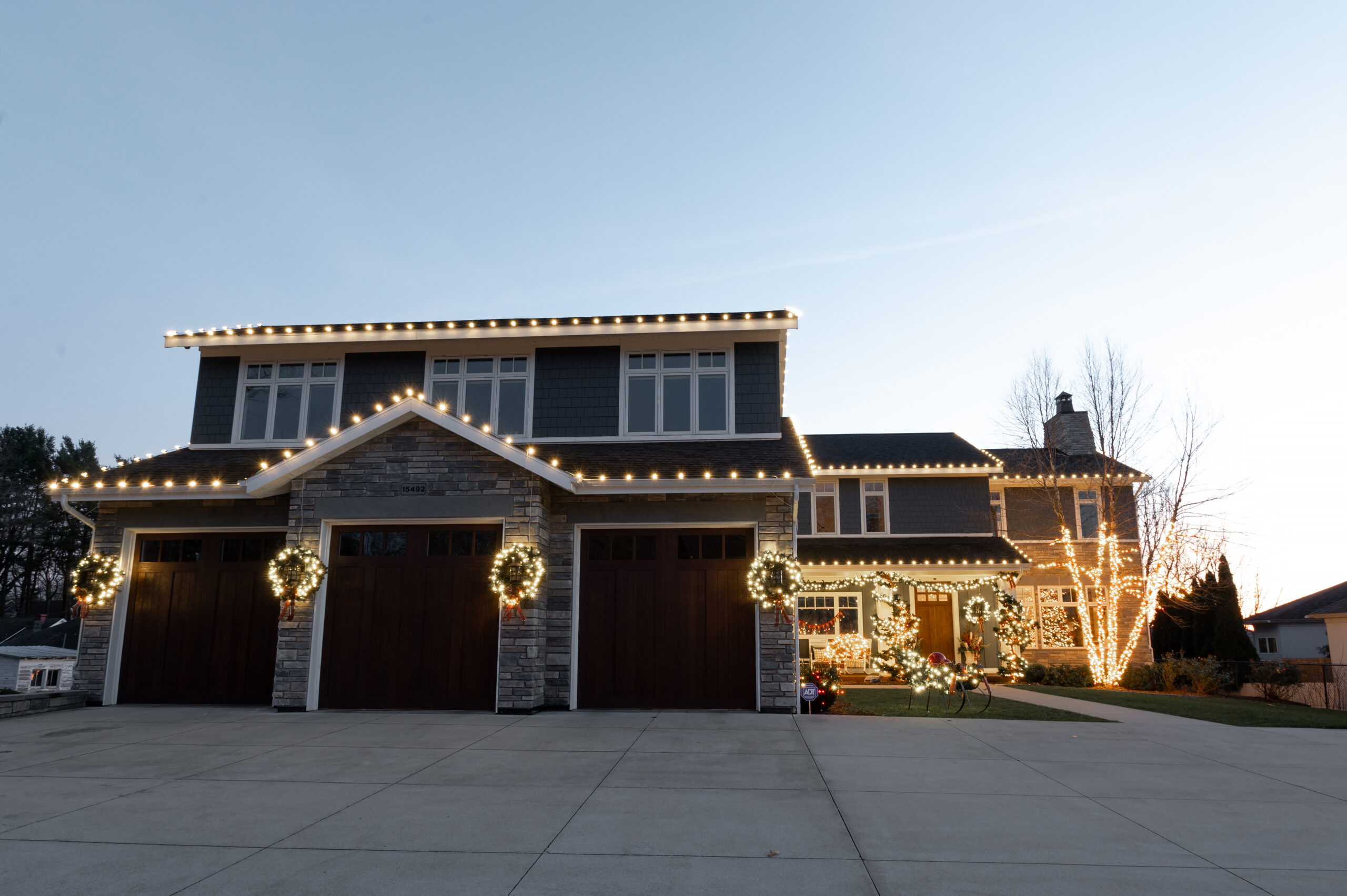 A large house is festively decorated with warm white Christmas lights outlining its roof, gables