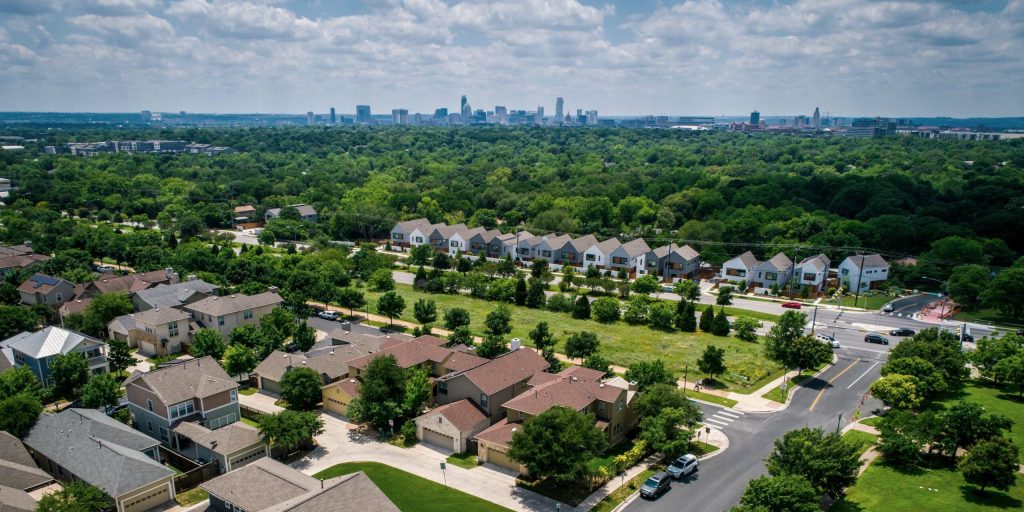An aerial view captures a residential neighborhood, lush green forest, and a distant city skyline under