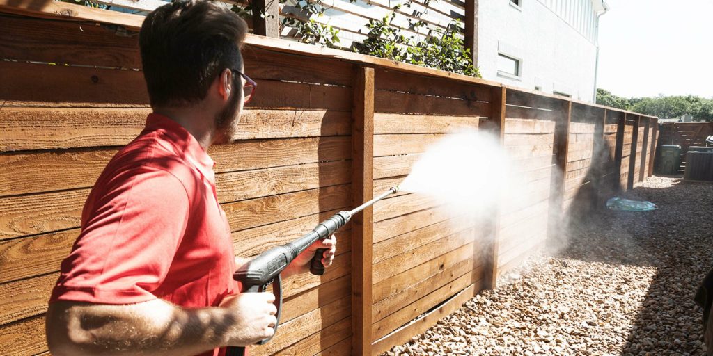 A man in a red shirt pressure washes a wooden fence, creating a cloud of water spray.