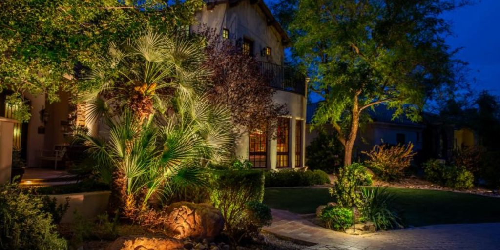 A stucco house and its lush garden are beautifully illuminated by warm landscape lighting at dusk.