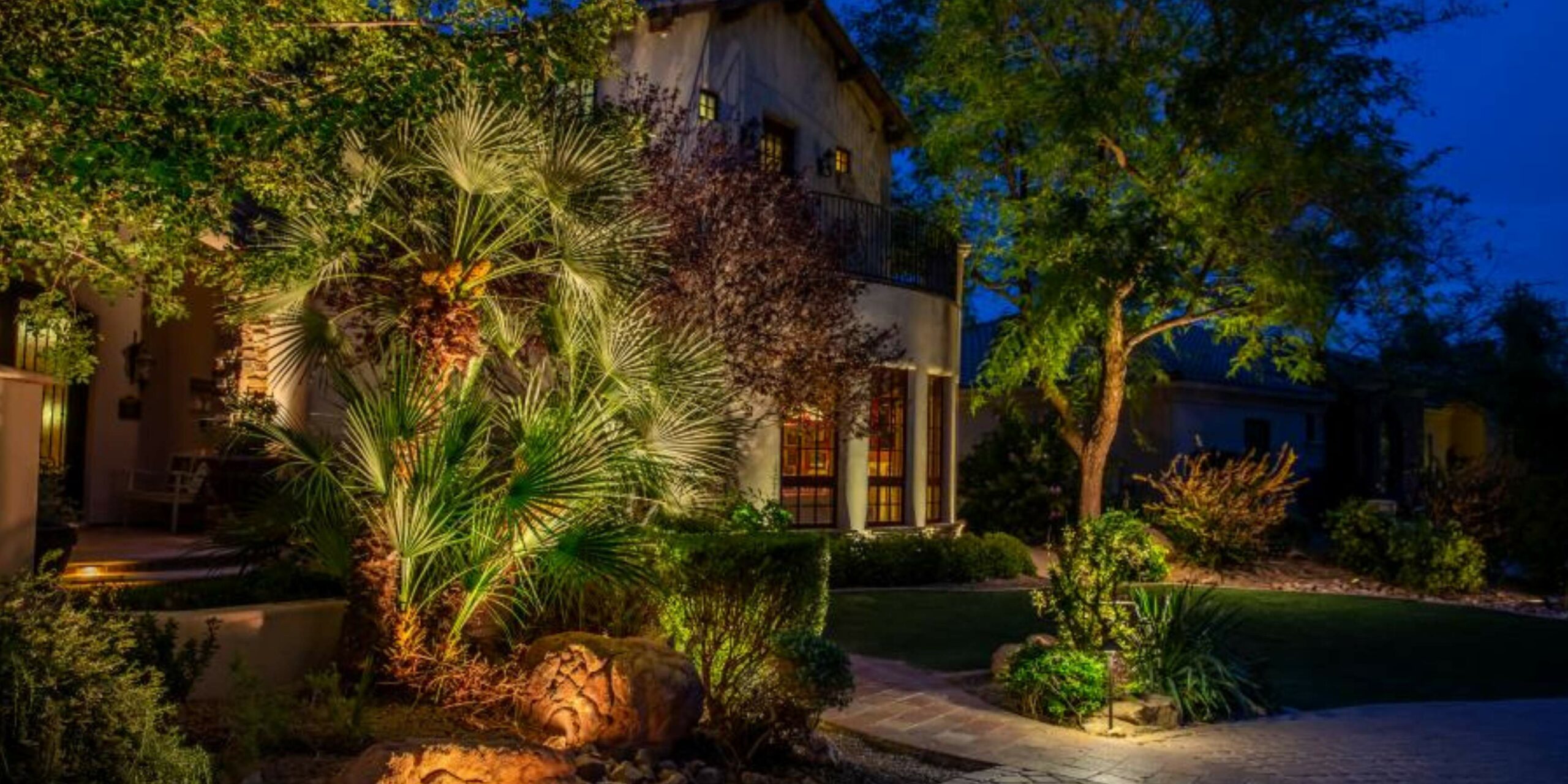 A stucco house and its lush garden are beautifully illuminated by warm landscape lighting at dusk.