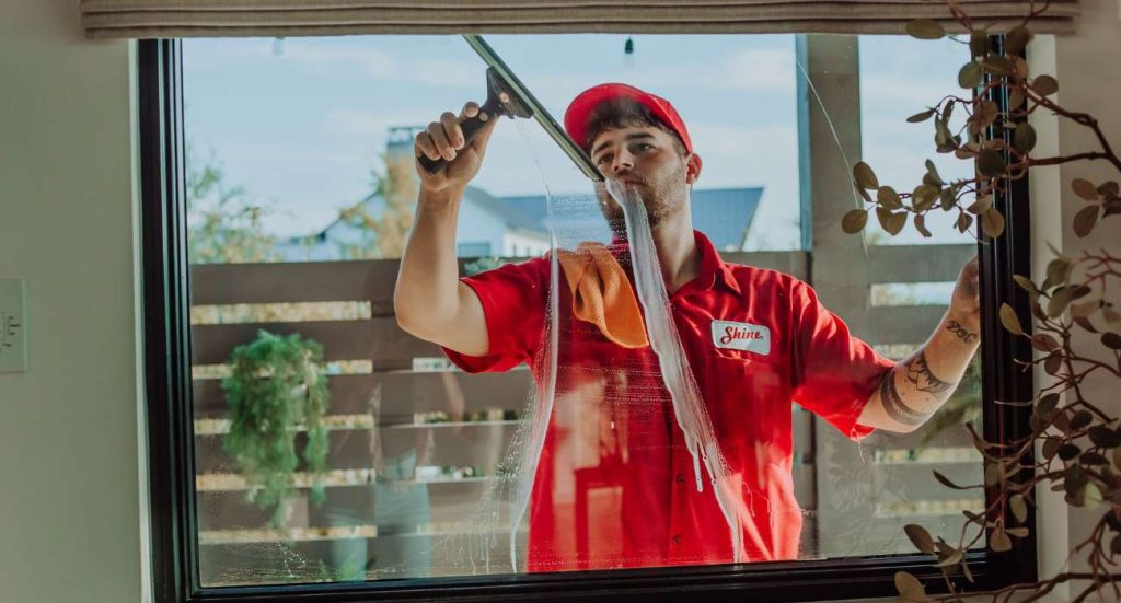 A man in a red uniform with a "Shine" logo expertly cleans a large window with