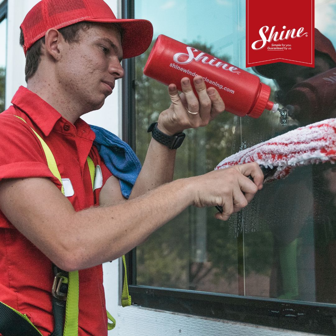 A professional window cleaner in a red uniform applies cleaning solution to a window.
