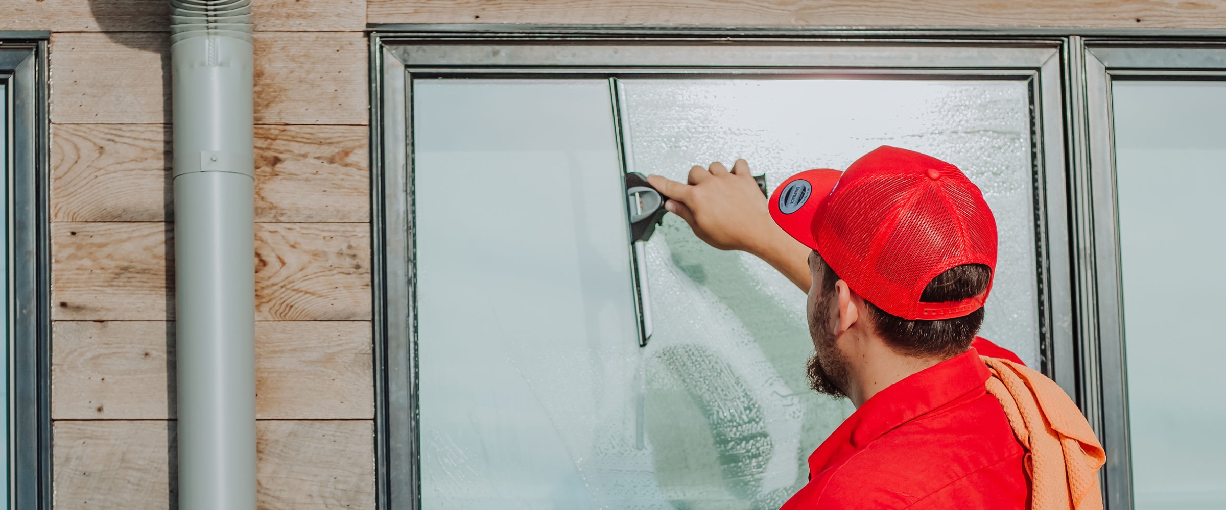 A person in a red cap cleans a window with a squeegee on a wooden-sided building.