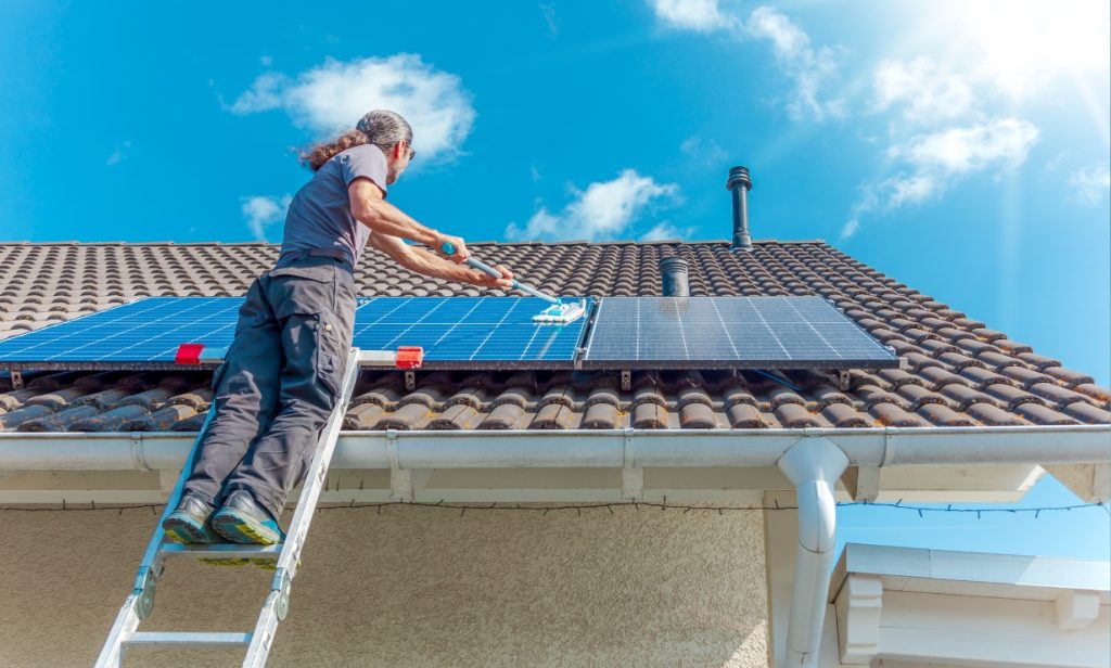 A person on a ladder cleans solar panels mounted on a tiled roof under a bright blue sky
