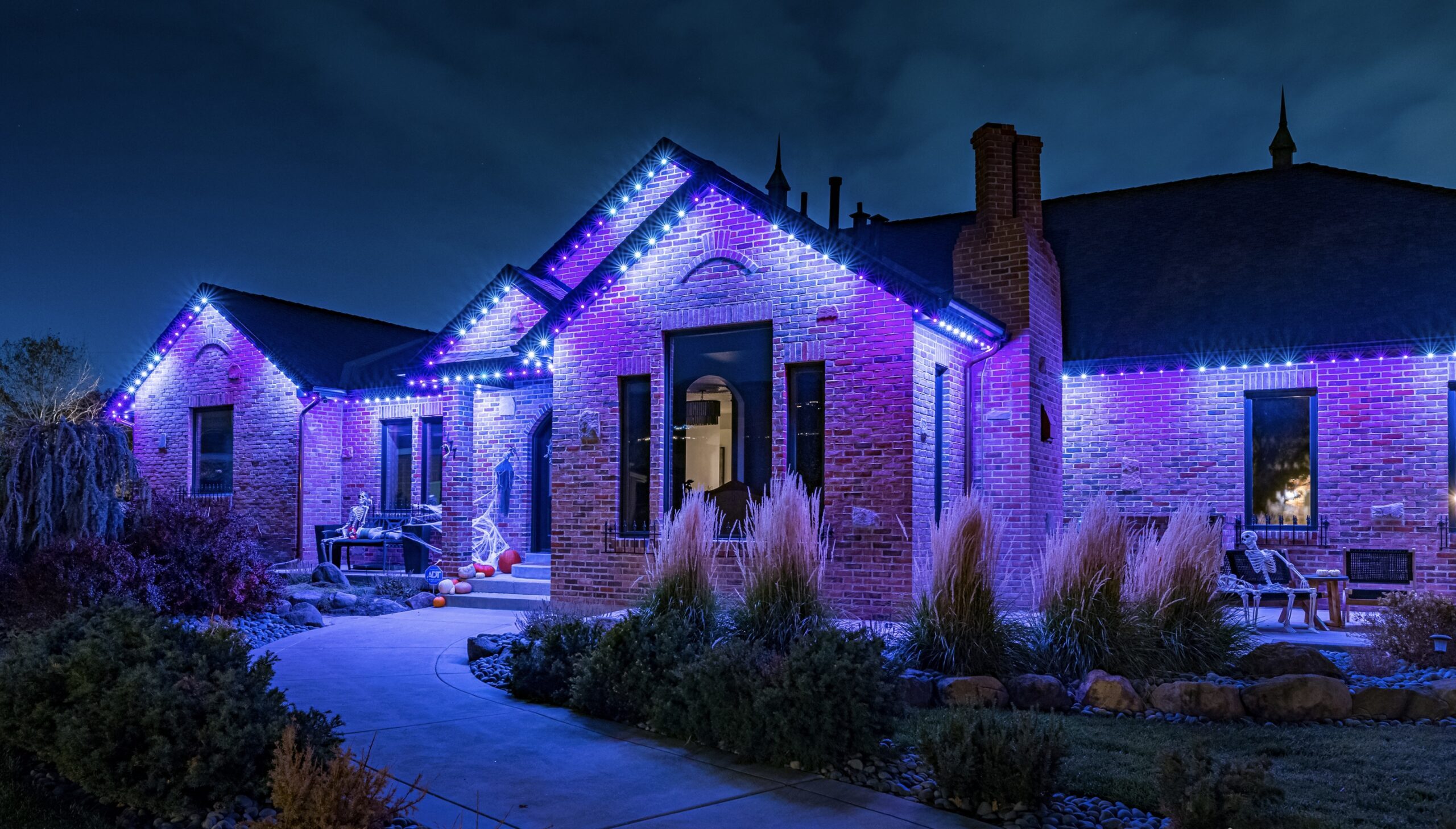 A brick house decorated for Halloween glows with vibrant purple and blue lights at night.