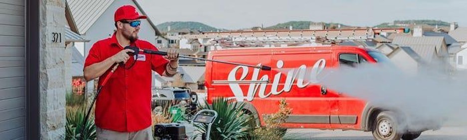 A professional in a red uniform pressure washes a surface with a red Shine service van nearby