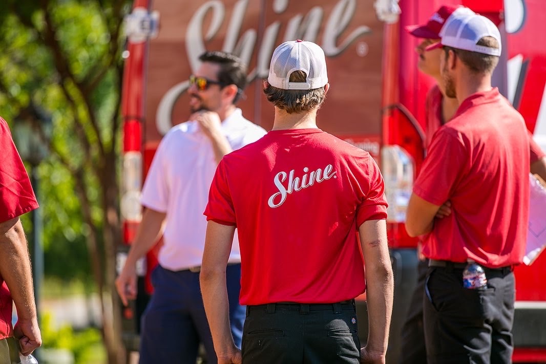 A person in a red "Shine" t-shirt and white cap stands with others