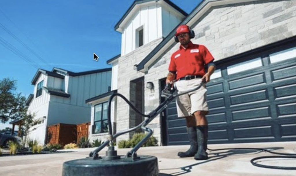 A professional in a red shirt uses a surface cleaner to power wash a concrete driveway.