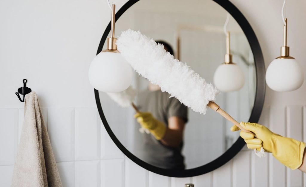 A yellow-gloved hand holds a white duster, reflected cleaning a round mirror in a