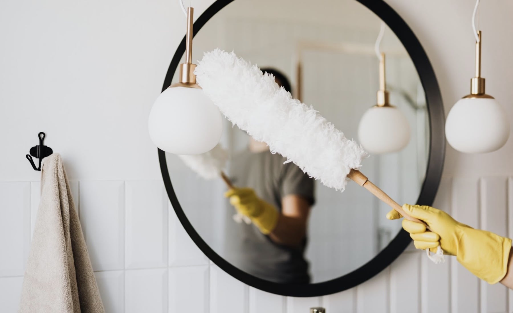 A yellow-gloved hand holds a white duster, reflected cleaning a round mirror in a
