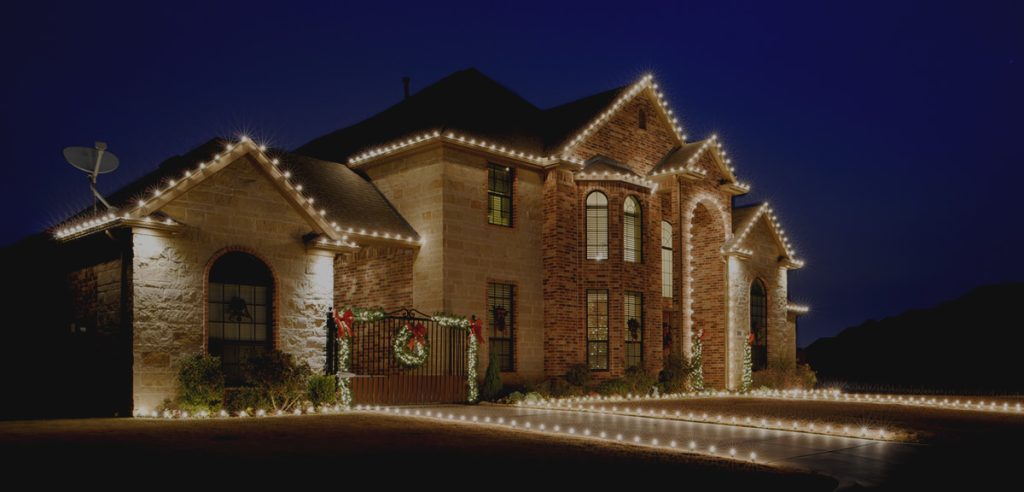 Elegant brick and stone house illuminated by sparkling white holiday lights along its roof and driveway. TITLE
