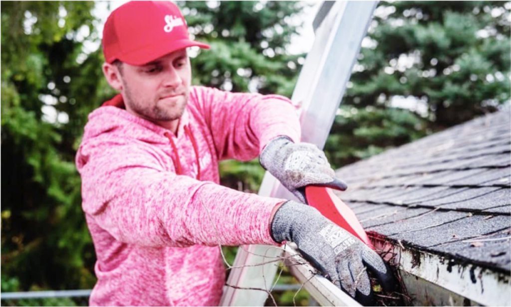 A gloved man on a ladder cleans a roof gutter filled with leaves using a red scoop tool