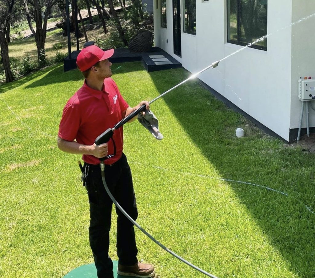 A man in a red uniform pressure washes the side of a white building on a sunny day.