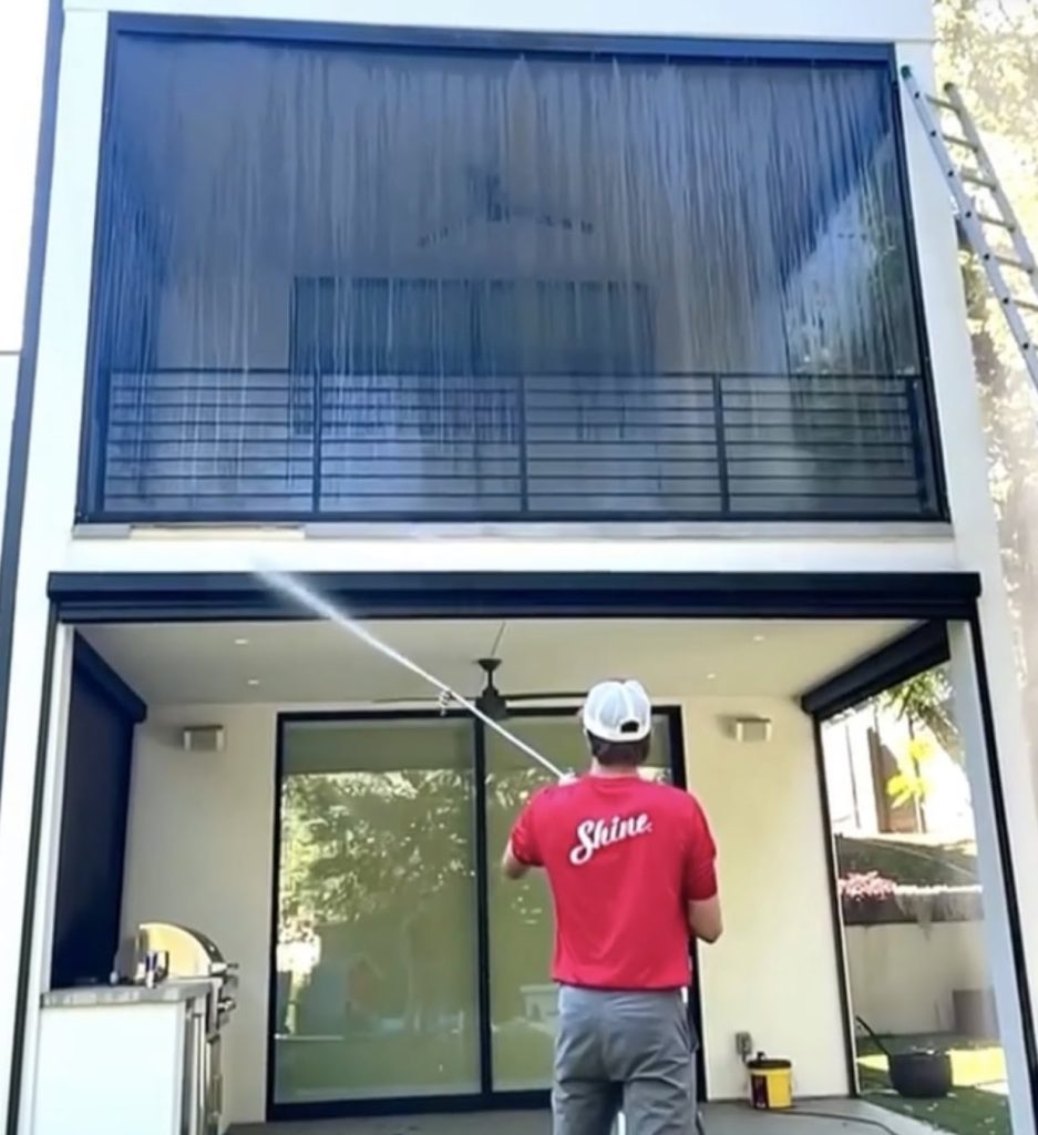 A man in a red 'Shine' shirt power washes the large glass panels of a