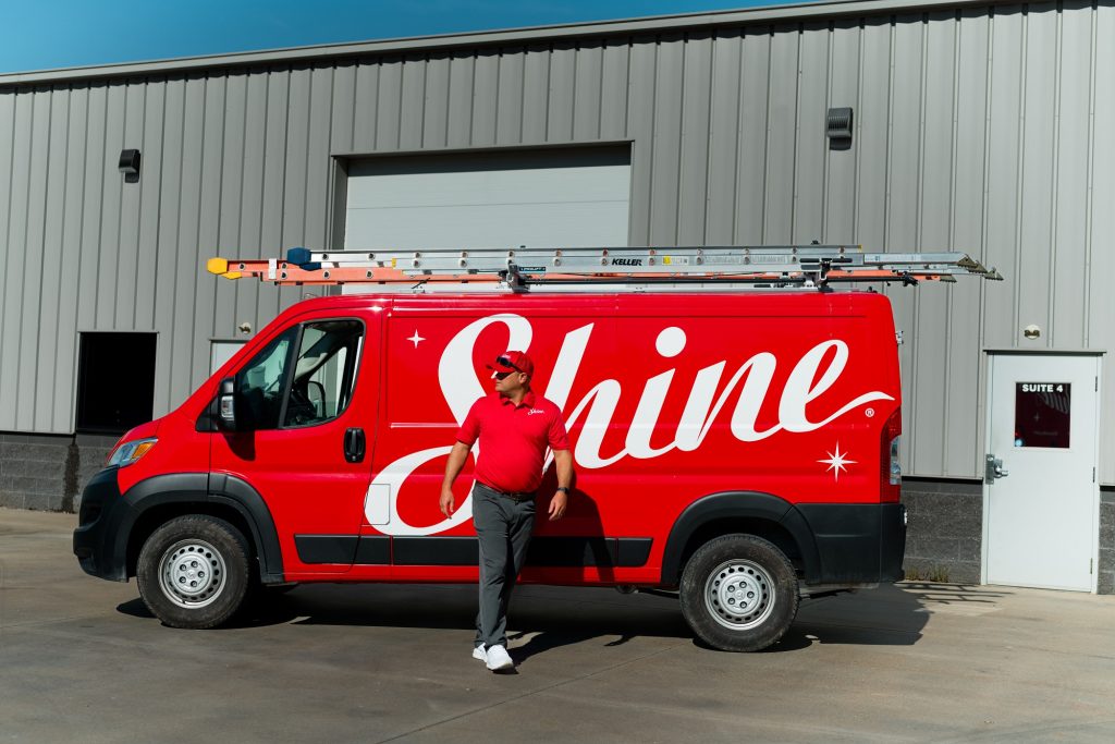 A technician in a red uniform stands by a red Shine service van with a roof ladder.