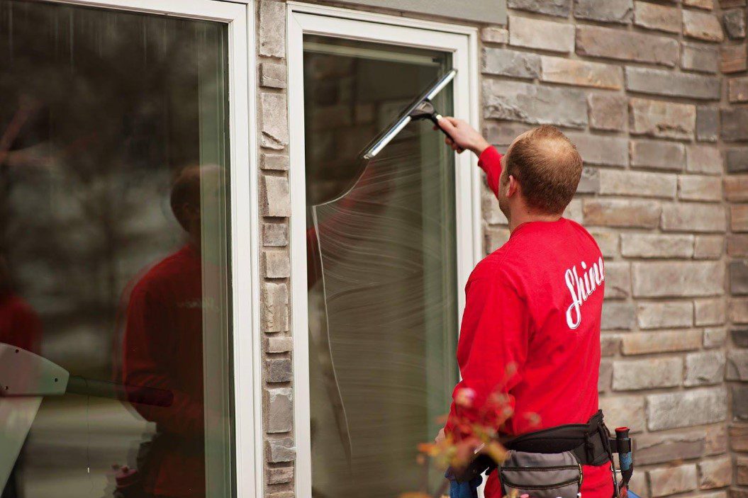 A service professional wearing a red 'Shine' shirt squeegees a residential window.