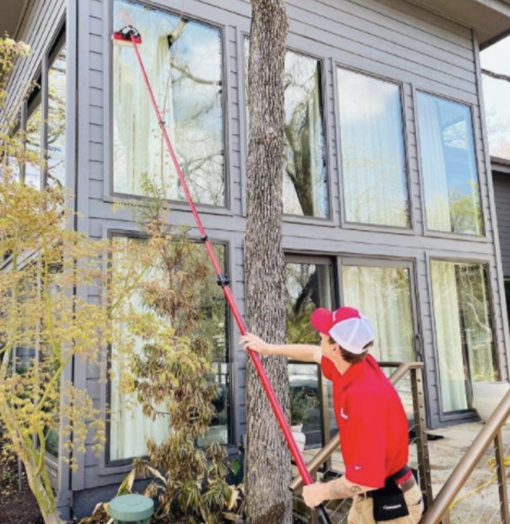 A person in a red uniform uses a long pole to clean a tall window on a modern grey