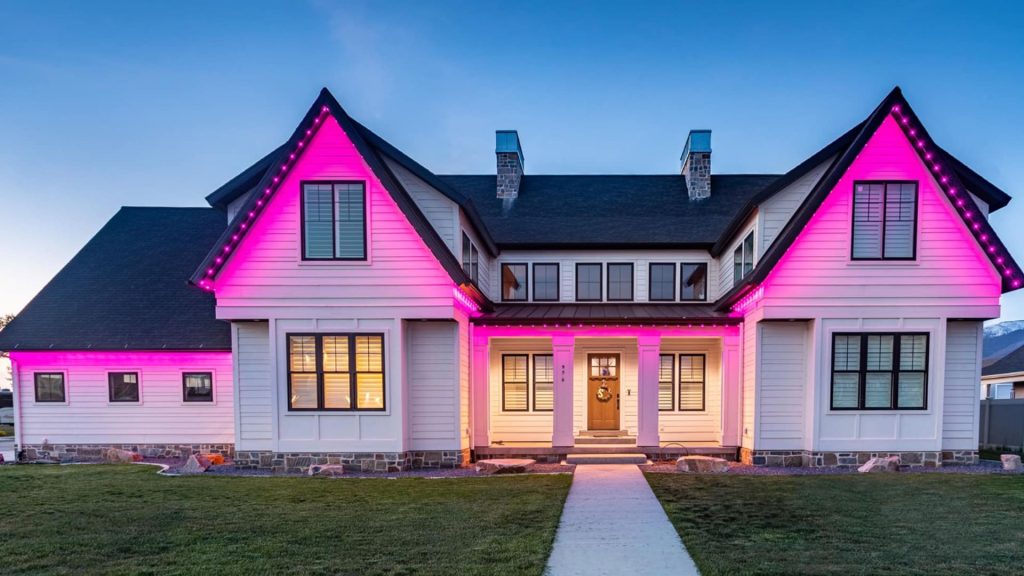 A modern white farmhouse with dark roofs is illuminated by bright pink lights at dusk.