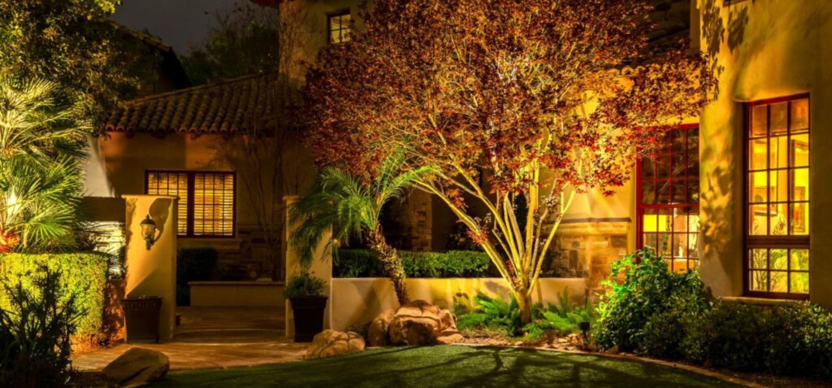 Warm landscape lighting illuminates a house exterior at night, highlighting a tree with red leaves