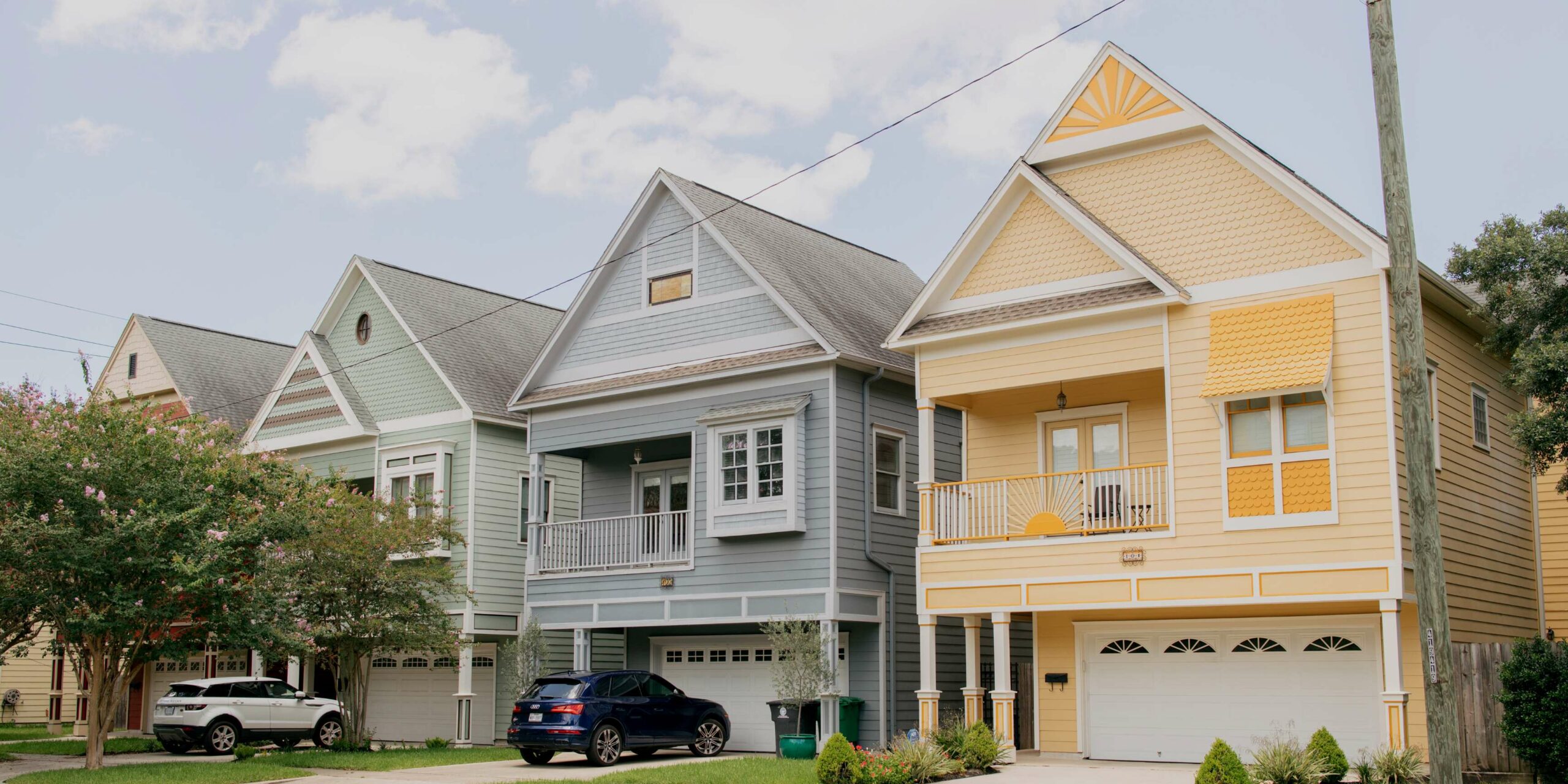 Four pastel-colored elevated houses with ground-level garages line a street with cars and