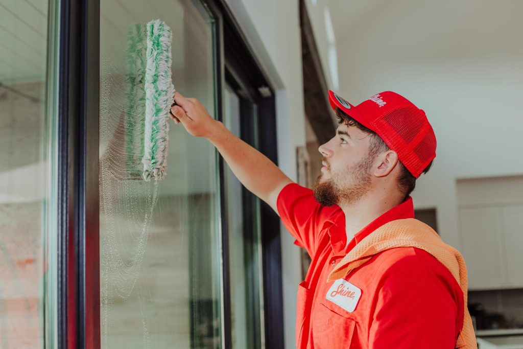 A professional window cleaner in a red uniform washes a large glass window with a fuzzy applicator.