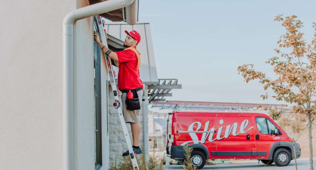A worker in a red uniform on a ladder services a building with a Shine van nearby.