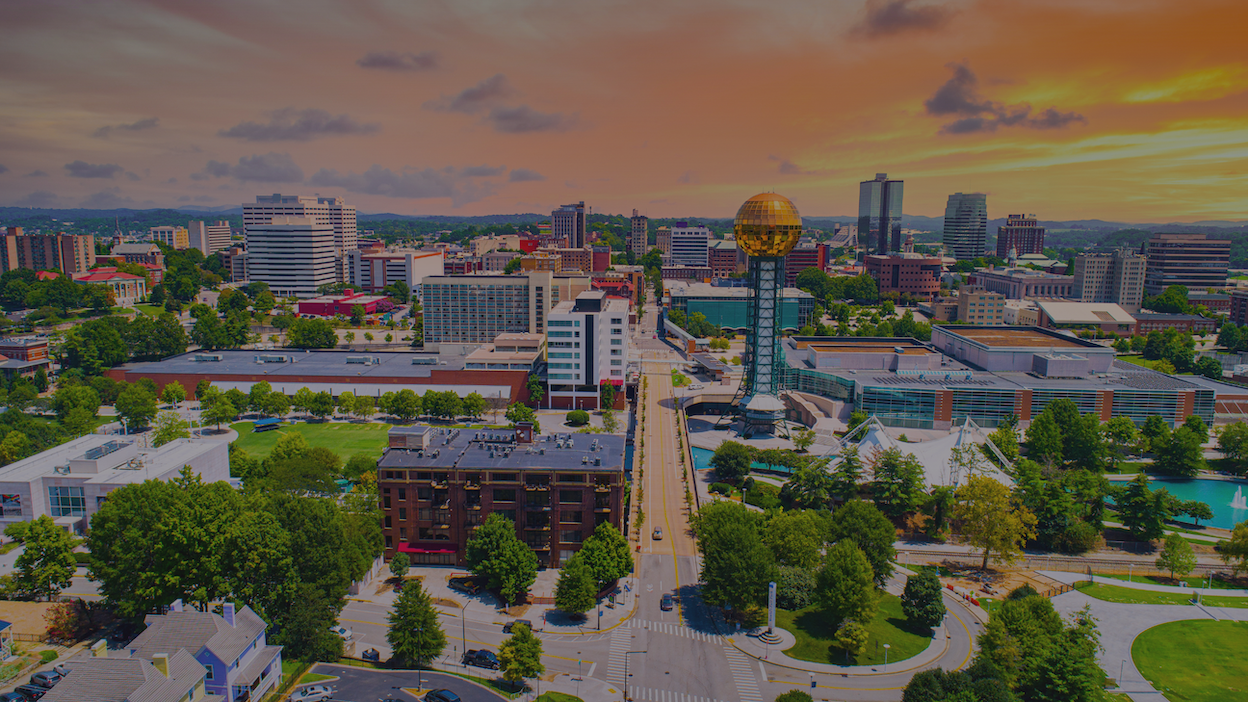 Aerial view of Knoxville, Tennessee skyline featuring the Sunsphere at sunset, surrounded by buildings and green trees.