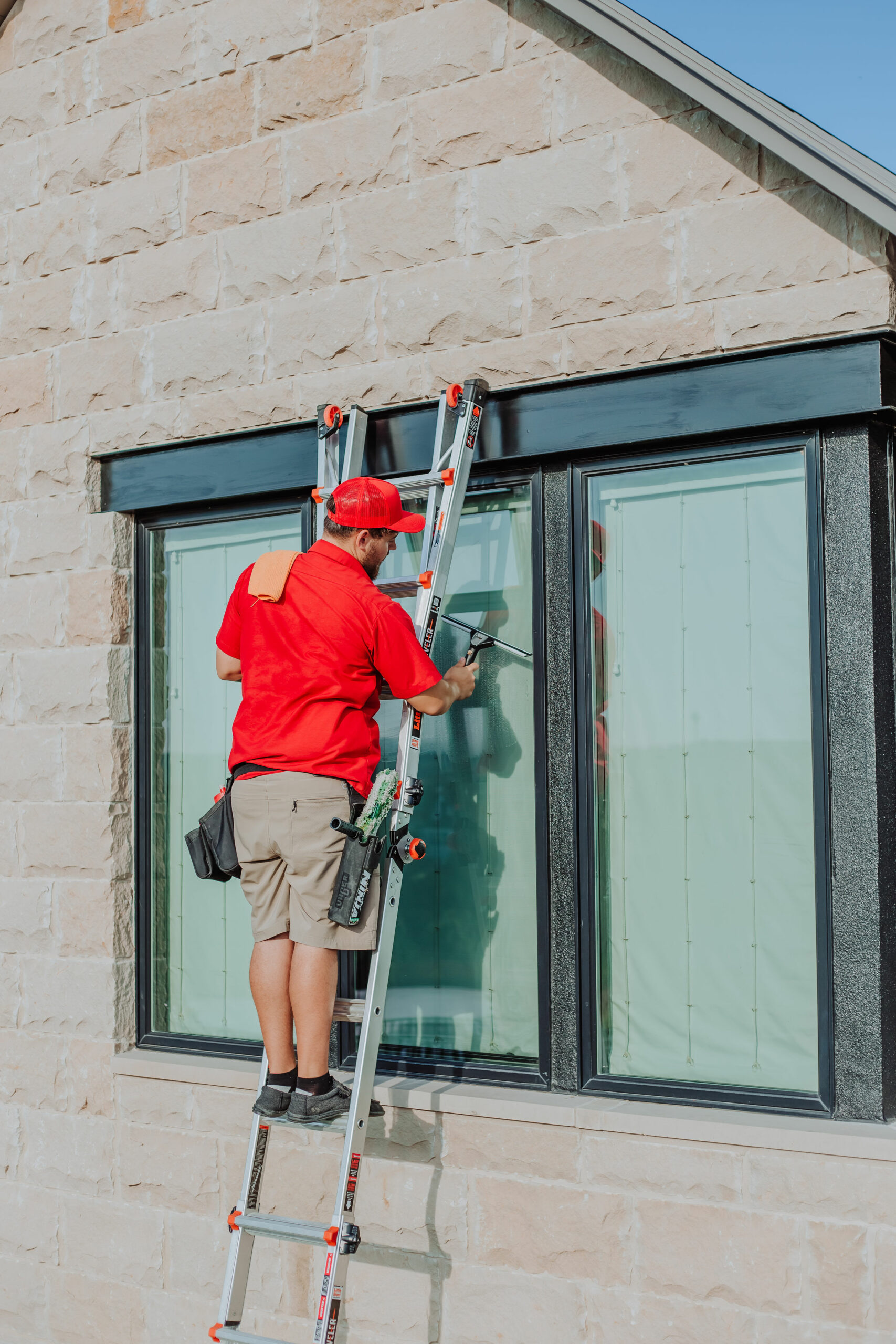 A man on a ladder cleans a large window on a stone building with a squeegee. TITLE