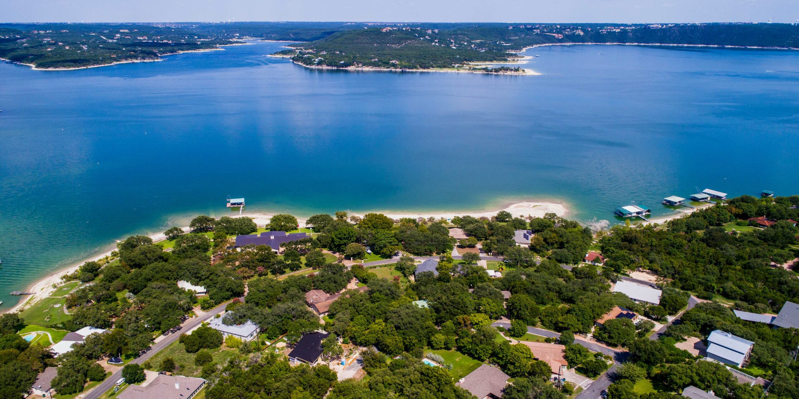 Aerial view of a vibrant blue lake with a tree-covered shoreline featuring numerous houses and boat docks.