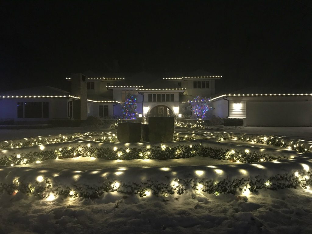 Snow-covered house and yard glow with festive warm white and colorful holiday lights at night.