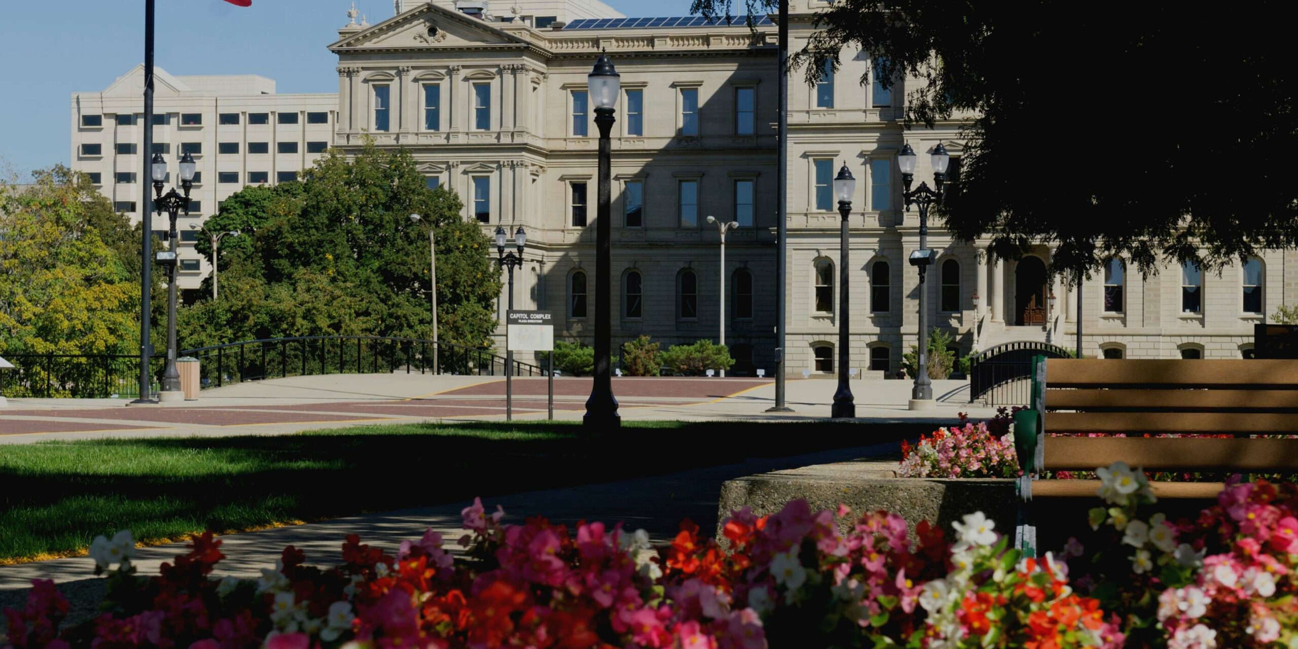 Lush flowers and a park bench frame a historic capitol complex building under a clear sky.