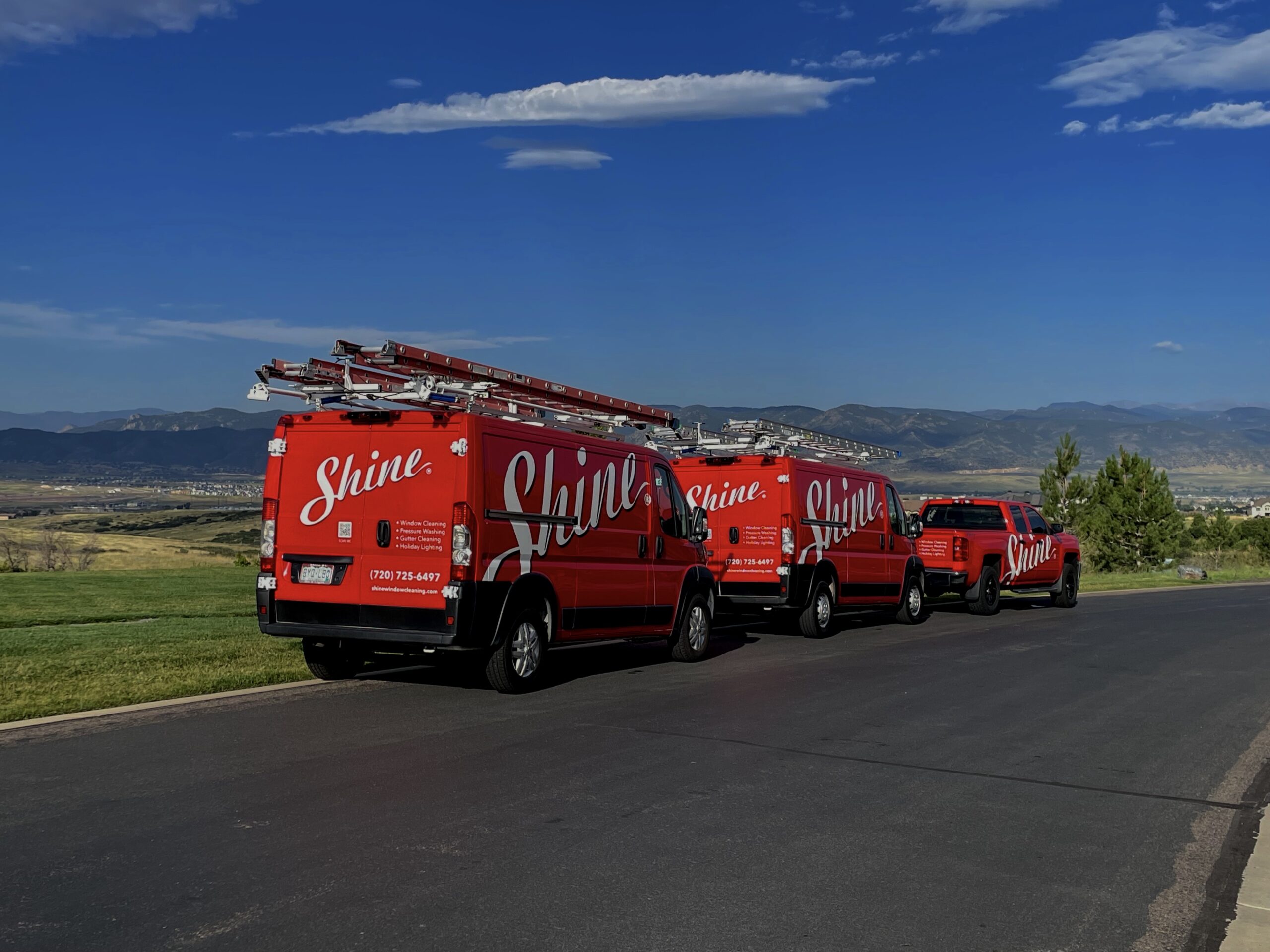 Red Shine service vans and truck with ladders parked on a road overlooking a mountain valley.