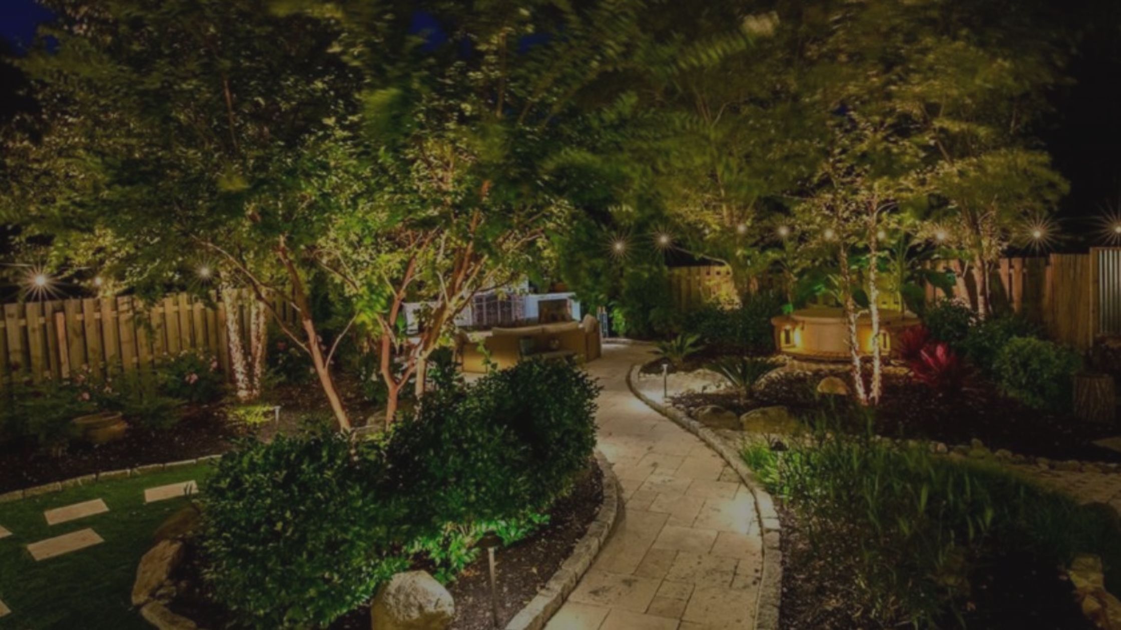 Lush backyard landscaping with a winding illuminated path and outdoor seating at night. TITLE: